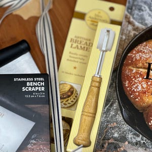 May include: A bread-making kit featuring a stainless steel bench scraper (6 in x 3 in), a wooden-handled bread lame, and a striped ribbon. The kit includes a box with the text "STAINLESS STEEL BENCH SCRAPER" and a package labeled "ARTISAN BREAD LAME".
