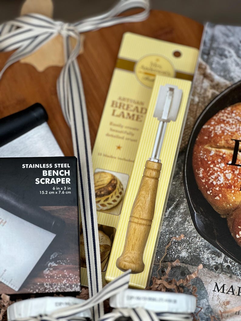 May include: A bread-making kit featuring a stainless steel bench scraper (6 in x 3 in), a wooden-handled bread lame, and a striped ribbon. The kit includes a box with the text "STAINLESS STEEL BENCH SCRAPER" and a package labeled "ARTISAN BREAD LAME".