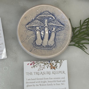 May include: A small, white ceramic dish with a blue and white mushroom design. The dish is hand-formed and decorated with food-safe glazes. The text "THE TREASURE KEEPER" is printed on a card next to the dish. The card reads: "I am hand formed from fine ceramic and decorated with bright, beautiful food safe glazes by the Walton family in Star, NC. Leave it to me to keep your items so you'll always remember where you left them. From clay to glazes to the box that holds me, I'm locally sourced and ready to keep all treasures at hand."