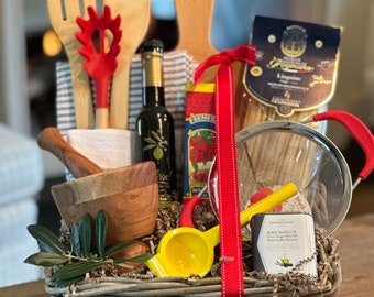 May include: A wicker basket filled with Italian-themed kitchen items, including a wooden mortar and pestle, a yellow citrus juicer, a red silicone spatula, wooden spoons, a bottle of olive oil, a jar of black truffle oil, a package of pasta, and a red and white striped kitchen towel.