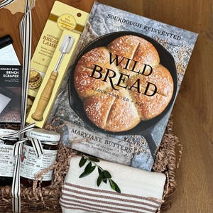 May include: A gift basket with a wooden cutting board, a cookbook titled "Wild Bread", and a bench scraper. The basket also contains a jar of jam, a bread lame, and a striped towel. The cookbook features a photo of baked bread.