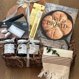 May include: A bread-making gift basket with a wooden cutting board, a cookbook titled "Wild Bread," and various baking tools. The basket includes jars of preserves, a bench scraper, and a striped towel with fringe.