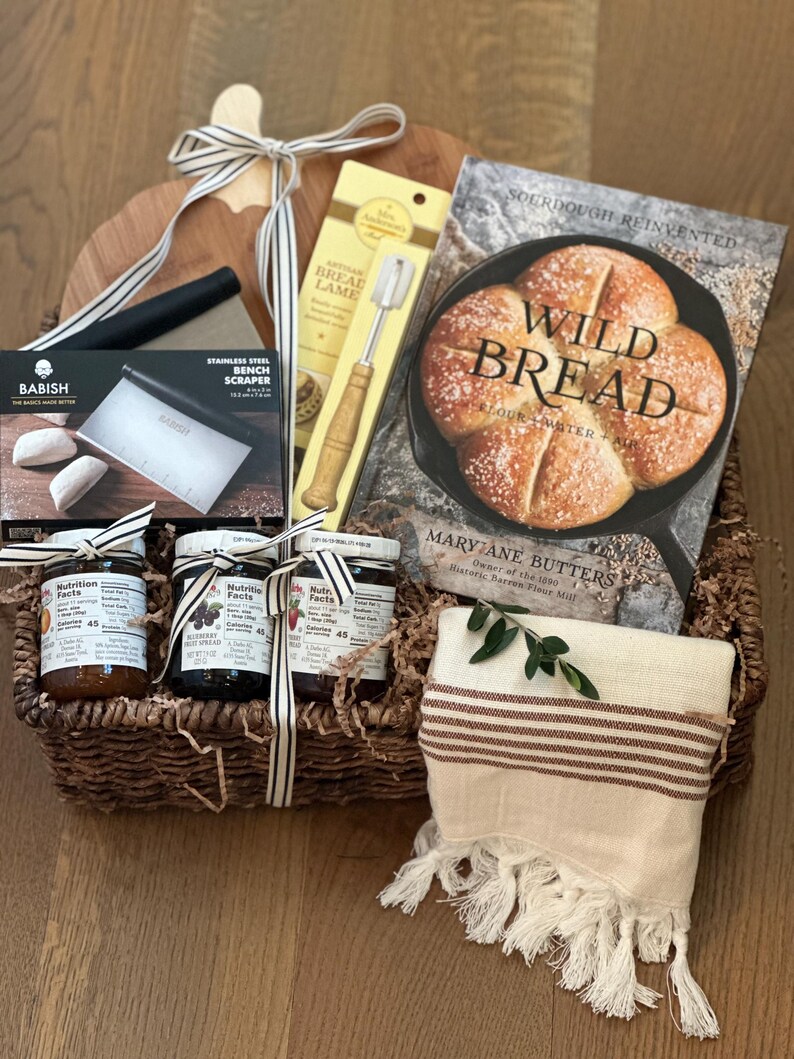 May include: A bread-making gift basket with a wooden cutting board, a cookbook titled "Wild Bread," and various baking tools. The basket includes jars of preserves, a bench scraper, and a striped towel with fringe.