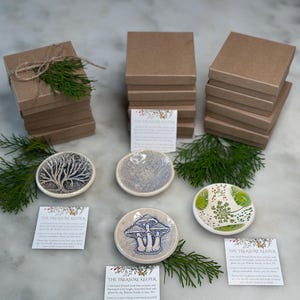 May include: Set of four small ceramic trinket dishes with various designs, including a tree, mushrooms, and flowers. Each dish comes with a small card that reads "The Treasure Keeper" and a description of the dish. The dishes are stacked on top of brown boxes.
