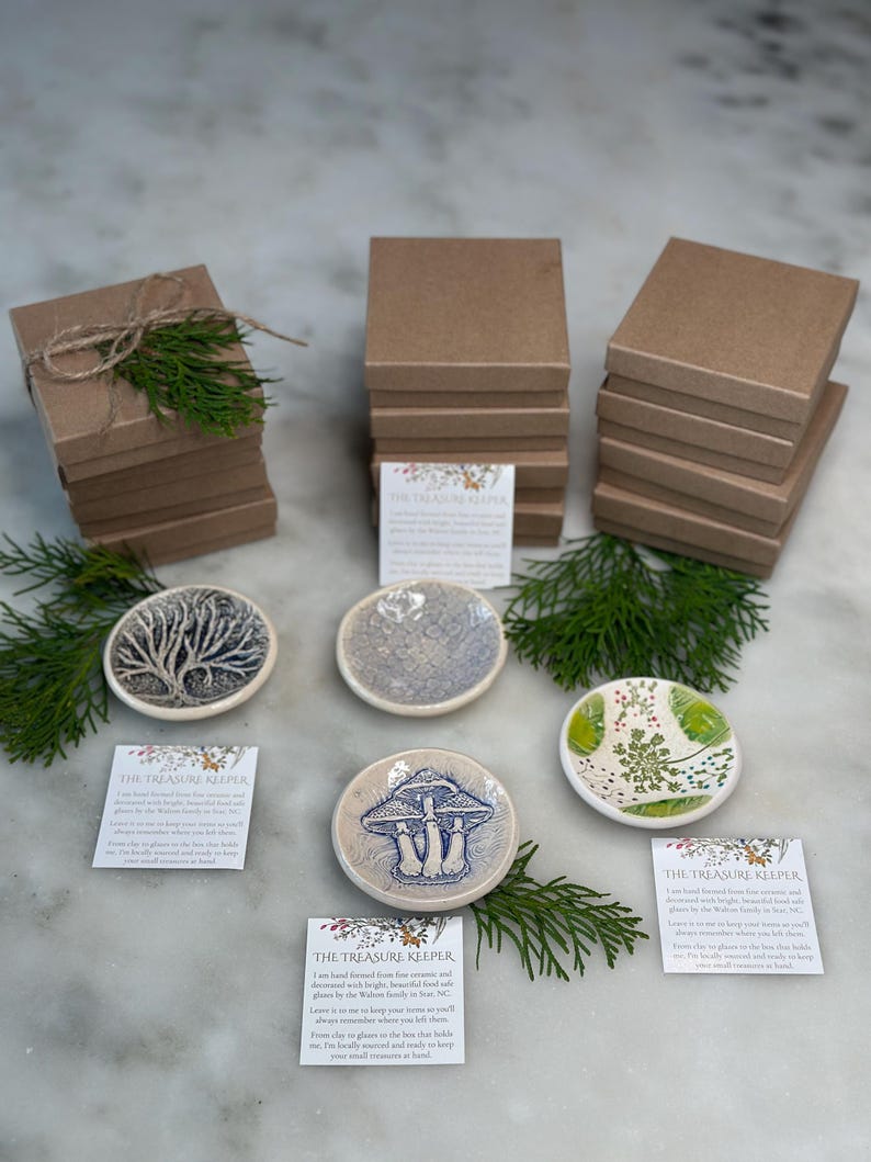 May include: Set of four small ceramic trinket dishes with various designs, including a tree, mushrooms, and flowers. Each dish comes with a small card that reads "The Treasure Keeper" and a description of the dish. The dishes are stacked on top of brown boxes.