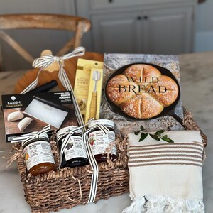 May include: A wicker basket filled with baking and kitchen items. Includes a wooden cutting board, a bread lame, a bench scraper, jars of fruit spread, a cookbook titled "Wild Bread", and a striped towel. The basket is tied with a striped ribbon.