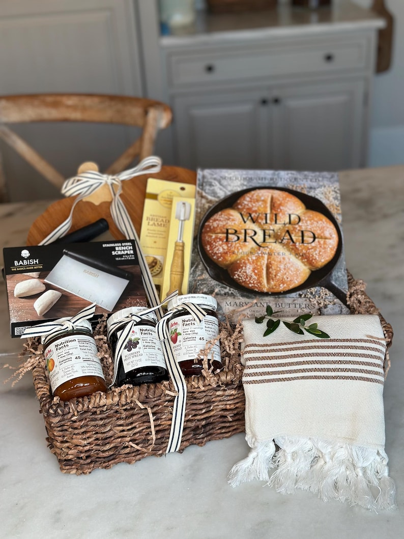 May include: A wicker basket filled with baking and kitchen items. Includes a wooden cutting board, a bread lame, a bench scraper, jars of fruit spread, a cookbook titled "Wild Bread", and a striped towel. The basket is tied with a striped ribbon.