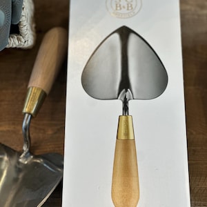 May include: A heart-shaped hand trowel with a wooden handle and a brass ferrule. The trowel is packaged in a white box with the text "S.C. B+B" printed on the front.