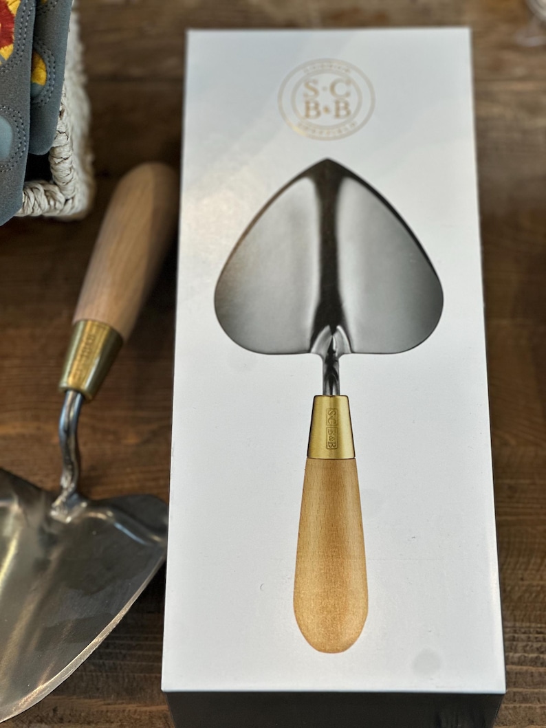 May include: A heart-shaped hand trowel with a wooden handle and a brass ferrule. The trowel is packaged in a white box with the text "S.C. B+B" printed on the front.
