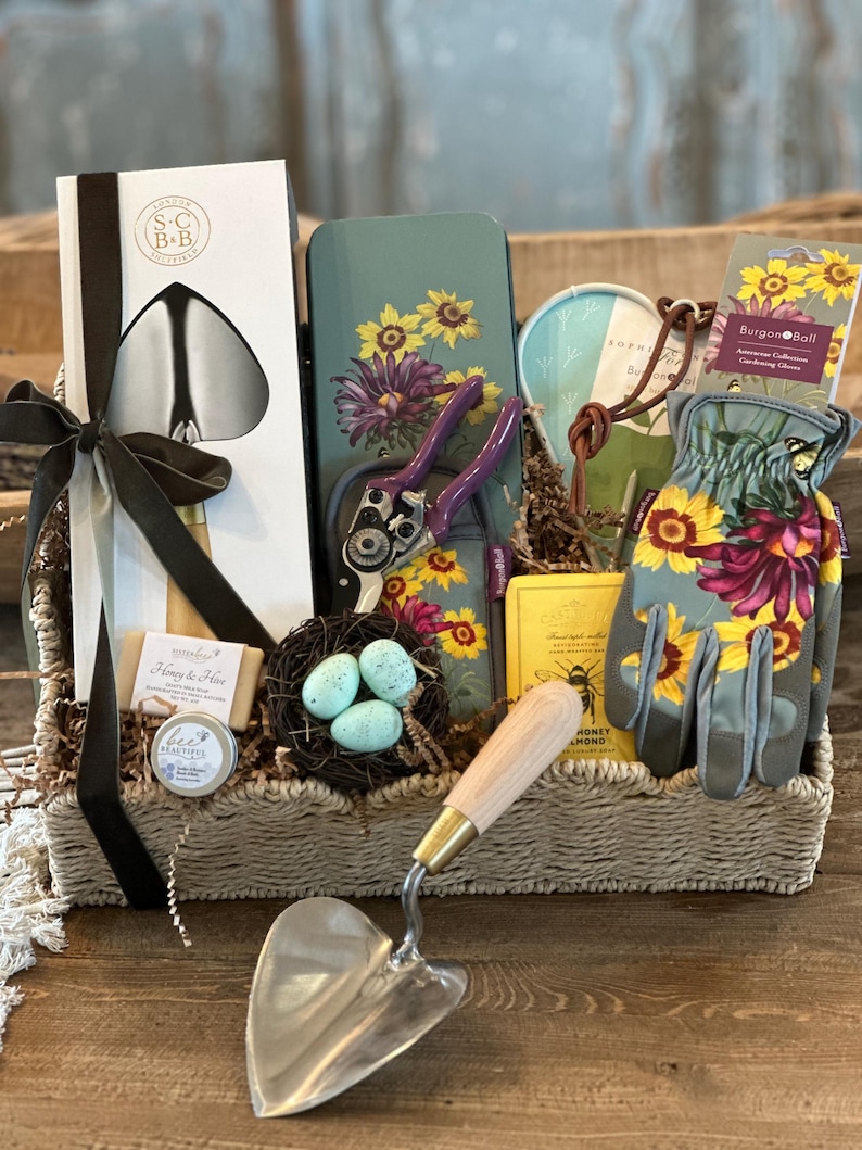 May include: A wicker basket filled with gardening themed gifts. The basket includes a pair of gardening gloves, a trowel, a pair of pruning shears, a small tin with a floral design, a bar of soap, a small jar of hand cream, a small bird's nest with blue eggs, and a book titled "Honey & Hive".