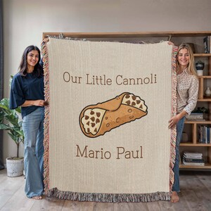 May include: A beige woven throw blanket featuring a cartoon cannoli illustration with the text "Our Little Cannoli" and "Mario Paul" in brown. The blanket has a fringed edge with pink, orange, and beige threads.