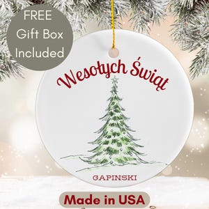 May include: White ceramic Christmas ornament with a green illustrated Christmas tree and the text "Wesołych Świąt" in red. The ornament also includes the text "GAPINSKI" and "Made in USA". A gold string is attached for hanging.