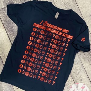 May include: A navy blue t-shirt with a red graphic of the Avengers logo and the text "I AVENGED MY WAY THROUGH 100 DAYS OF SCHOOL". The graphic is a repeating pattern of the Avengers characters' symbols.