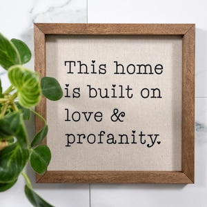 May include: Square wooden framed sign with the text "This home is built on love & profanity." The sign has a neutral canvas background and black lettering. A green leafy plant is in the upper left corner.