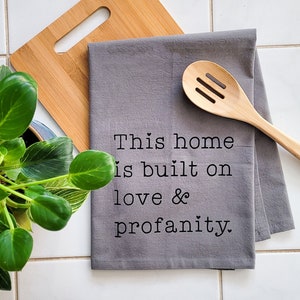 May include: A gray kitchen towel with the text "This home is built on love & profanity."  The towel is folded and lying on a white tiled surface. A wooden spoon and a wooden cutting board are also in the image.