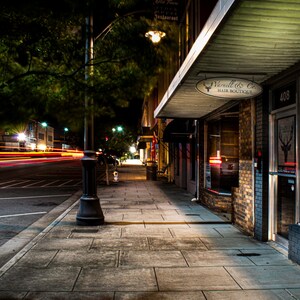 May include: Nighttime street scene with a sidewalk, streetlights, and storefronts. The storefront on the right is a hair boutique with a sign that reads "Vanell & Co. Hair Boutique." The street has streaks of light from passing vehicles.