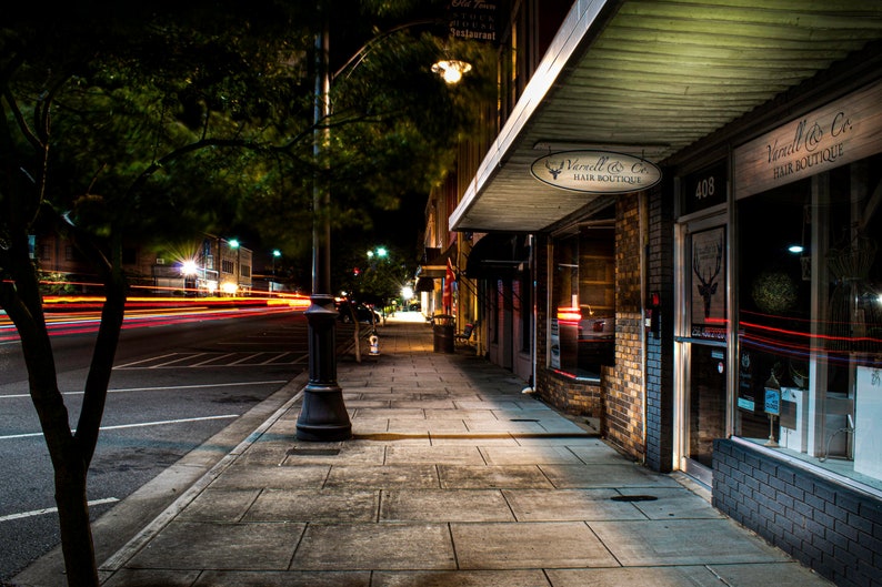 May include: Nighttime street scene with a sidewalk, streetlights, and storefronts. The storefront on the right is a hair boutique with a sign that reads "Vanell & Co. Hair Boutique." The street has streaks of light from passing vehicles.