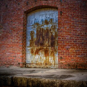 May include: A weathered metal door with rust-colored streaks is set into a red brick wall. The door is arched at the top and has a rectangular shape. The brick wall is made of red bricks.
