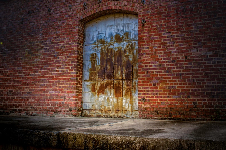 May include: A weathered metal door with rust-colored streaks is set into a red brick wall. The door is arched at the top and has a rectangular shape. The brick wall is made of red bricks.