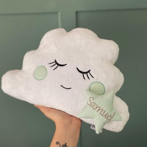 Può includere: Un cuscino a forma di nuvola bianca con occhi chiusi ricamati, ciglia e una bocca sorridente. È attaccata una stella verde chiaro con il nome "Samuel". Il cuscino ha una consistenza morbida e soffice.