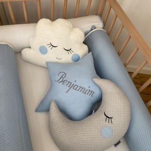Può includere: Un set di biancheria da letto per bambini in una culla. Include un cuscino a forma di nuvola bianca con dettagli blu, un cuscino a forma di stella blu con il nome "Benjamin" ricamato e un cuscino a forma di luna grigia.