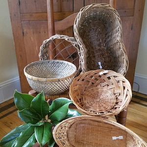 May include: Five woven baskets of various shapes and sizes, including a round basket, a rectangular basket, and an oval basket. The baskets are made of natural materials and have a rustic, handmade look. The baskets are arranged on a wooden chair with a green plant in the foreground.