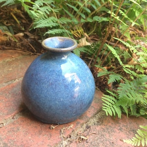 May include: A small, round, blue ceramic vase with a speckled glaze. The vase is sitting on a brick surface.
