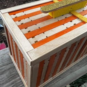 May include: A wooden box with orange and natural wood stripes. The box has a lid with a yellow and gold glitter object on top. The box is made of popsicle sticks and has a rustic, handmade appearance.