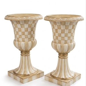 May include: Two decorative urns with a cream and gold color scheme. The urns have a checkered pattern on the body and a striped pattern on the base. The rims and accents are gold. The urns are on a square base.