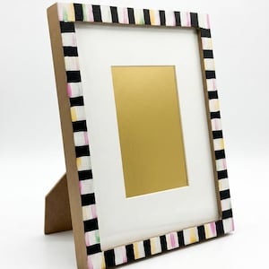 Pu&ograve; includere: Una cornice decorativa con un passe-partout bianco e un interno color oro. La cornice &egrave; dipinta con strisce bianche e nere, accentate con rosa, giallo e verde. La cornice &egrave; sostenuta da un supporto di cartone.