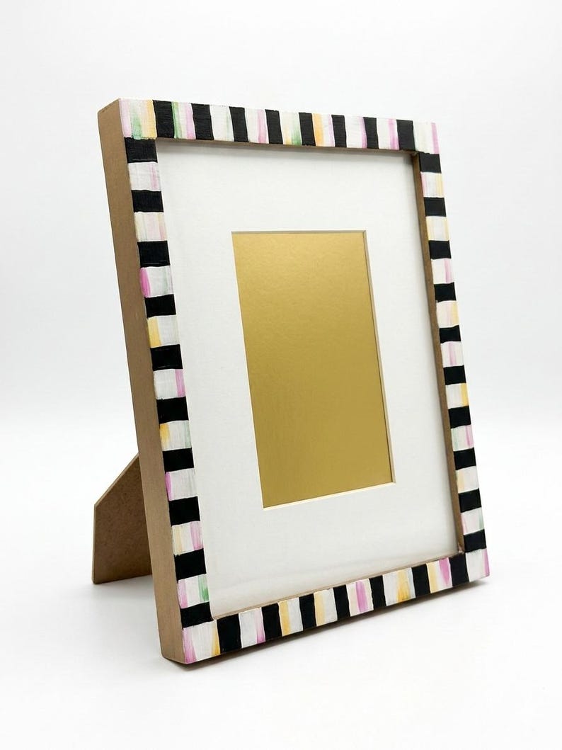 Pu&ograve; includere: Una cornice decorativa con un passe-partout bianco e un interno color oro. La cornice &egrave; dipinta con strisce bianche e nere, accentate con rosa, giallo e verde. La cornice &egrave; sostenuta da un supporto di cartone.