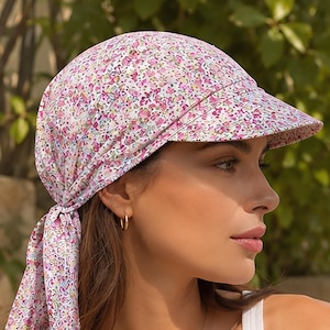 Puede incluir: Un pañuelo para la cabeza con visera rígida que presenta un denso estampado floral en tonos rosa, morado y blanco. La tela cubre la coronilla y se ata en la parte posterior con dos extremos sueltos, creando una silueta ajustada con visera.