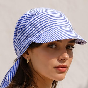 Puede incluir: Un pañuelo para la cabeza a rayas azules y blancas con una visera incorporada. La tela presenta una textura de seersucker con finas rayas verticales. El pañuelo está diseñado para atarse en la parte posterior, con largas tiras de tela.