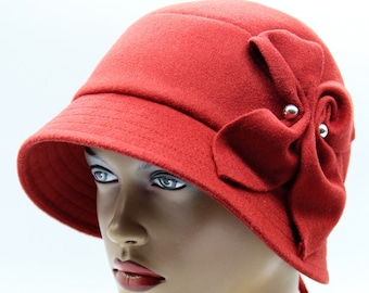 May include: A red cloche hat with a decorative bow on the side. The hat has a small brim and is made of a felt-like material. The bow is adorned with small, silver-colored embellishments. A ribbon detail hangs from the back of the hat.