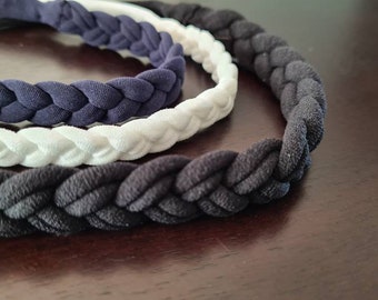 May include: Three braided headbands arranged in a semi-circle. The headbands feature a woven fabric texture and come in three distinct colors: a dark navy blue, a crisp white, and a solid black. Each band has a consistent braided pattern.