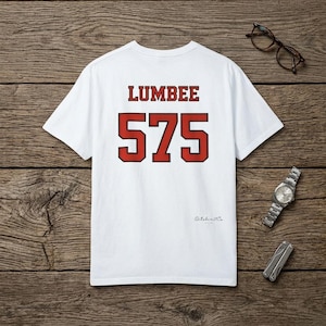 Könnte beinhalten: Weißes T-Shirt mit dem Wort "LUMBER" über der Zahl "575" in roten Blockbuchstaben. Das Shirt liegt auf einer Holzoberfläche mit Brille, Uhr und Multitool.