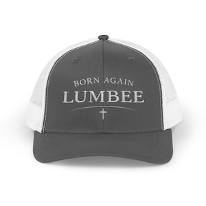 May include: A gray trucker hat with a white mesh back. The front of the hat features the text "BORN AGAIN LUMBEE" in a serif font, with a cross symbol below the text. The hat has a curved brim.