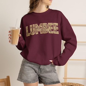 May include: A maroon long-sleeve crewneck sweatshirt featuring the word LUMBEE printed in a large, beige, block-letter font. The letters are filled with a detailed, intricate pattern of small flowers, leaves, and geometric shapes.