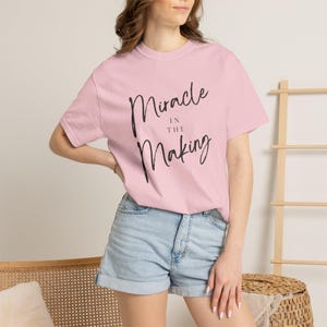 May include: A pale pink t-shirt featuring the text "Miracle in the Making" in black cursive script. The shirt is paired with denim shorts. A wooden ladder and a woven basket are visible in the background.