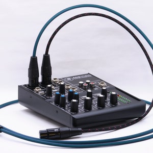 May include: A black audio mixer with multiple knobs and sliders, connected to two XLR cables with black connectors and teal and black braided sheathing. The mixer has the text "Mackie" on it.
