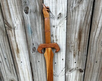 May include: A wooden sword with a dragon head pommel. The sword is brown and has a simple design. It is leaning against a wooden fence.