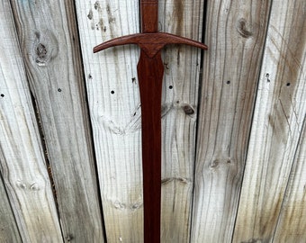 May include: A wooden sword with a carved wooden pommel. The sword is brown and has a simple design. The sword is hanging on a wooden fence.
