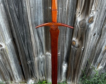 May include: A handcrafted wooden sword with a dark reddish-brown finish. The hilt features a carved design, and the blade has a long, slender profile. The sword is displayed against a weathered wooden fence and green grass.