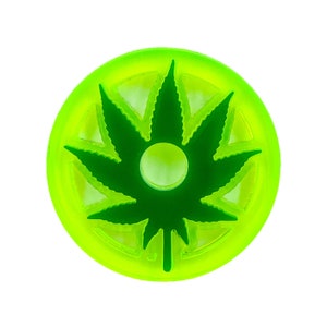 Puede incluir: Un disco redondo verde brillante con un diseño de hoja de marihuana verde oscuro. La hoja está recortada del disco, creando un agujero en el centro.