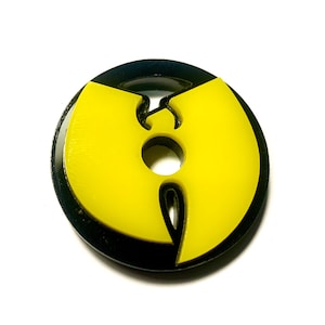 Puede incluir: Un amuleto con el logotipo de Wu-Tang Clan en negro y amarillo con un agujero en el centro.