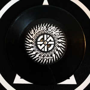 K&ouml;nnte beinhalten: Schwarze Schallplatte mit einem wei&szlig;en Label, das ein stilisiertes Logo mit dem Text "Lazer Life Los Angeles" und einem Blitzdesign zeigt.