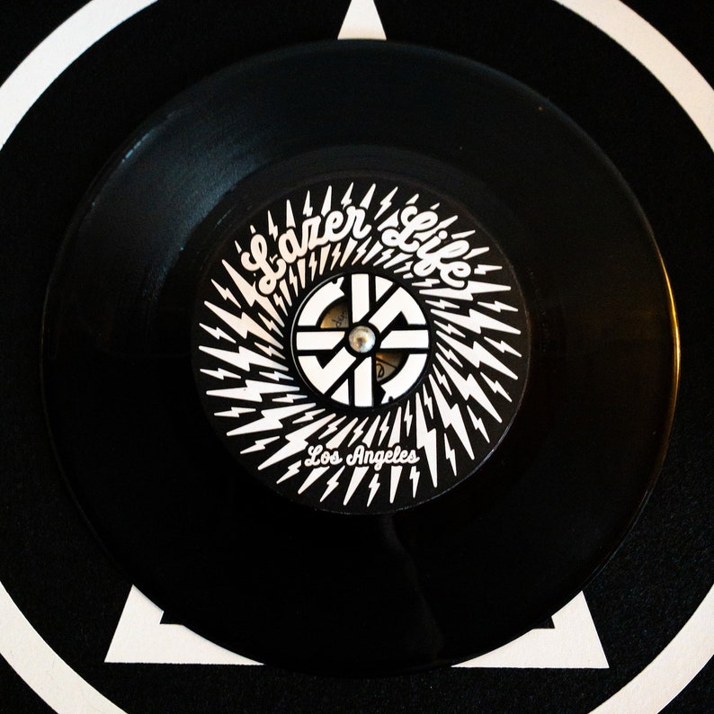 K&ouml;nnte beinhalten: Schwarze Schallplatte mit einem wei&szlig;en Label, das ein stilisiertes Logo mit dem Text "Lazer Life Los Angeles" und einem Blitzdesign zeigt.