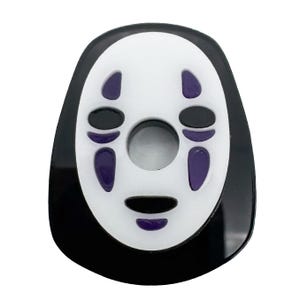 Może przedstawiać: Maska Bez Twarzy z filmu animowanego Spirited Away. Maska ma białą twarz z czarnymi otworami na oczy i usta oraz fioletowymi oznaczeniami. Maska ma czarną obwódkę.