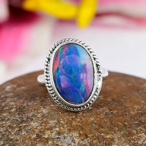May include: A silver ring with an oval-shaped opal gemstone. The opal displays a vibrant play of color, with hues of blue, purple, and red. The ring has a detailed rope-like border around the gemstone.