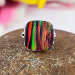 May include: A square-faced ring with a vibrant, multicolored opal stone. The opal displays streaks of red, green, and purple hues, set in a silver band. The ring is displayed on a textured, brown surface.